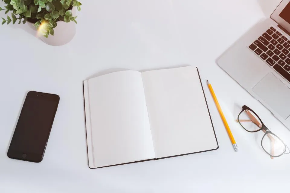 A phone, an open notebook, glasses and a laptop on a white desk