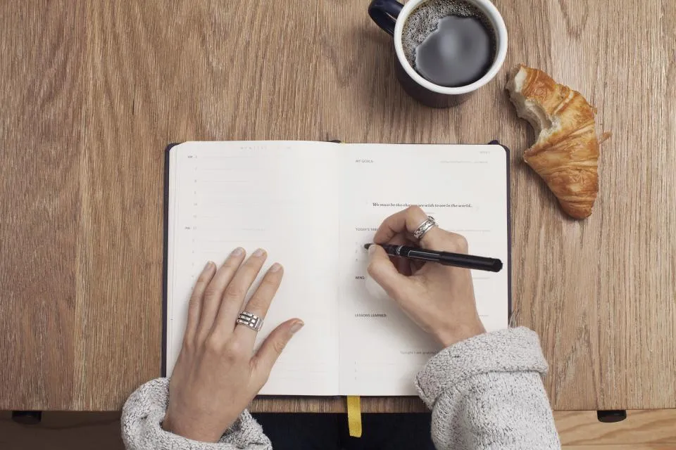 Hands writing in a journal next to a coffee and a croissant